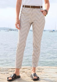 Beige gemusterte Hosen mit strukturiertem Design, ergänzt durch einen braunen Gürtel. Getragen mit schwarzen Sandalen auf einer steinigen Oberfläche in der Nähe von Wasser.