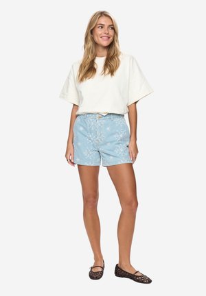 Kobieta stojąca, ubrana w oversize'ową białą koszulę z krótkim rękawem, jasnoniebieskie szorty z dżinsu w kwiatowy wzór oraz czarne plecione baleriny.