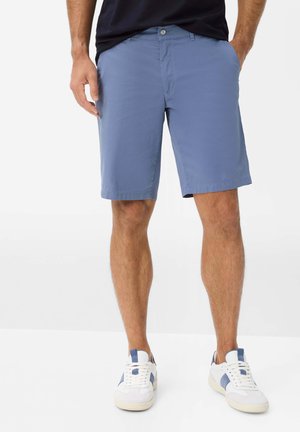 Homme portant un short bleu jusqu'au genou et des baskets blanches avec des accents bleus, debout avec les mains partiellement dans les poches.
