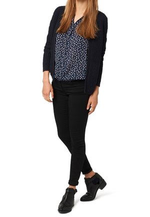 Black skinny jeans, navy knit cardigan, and patterned blouse featuring white flowers on a dark background. Black ankle boots with bow detail.