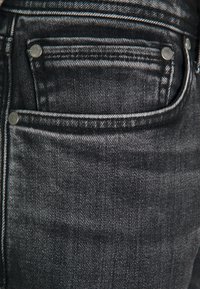 Černé džíny z denimu s mírným leskem, oblečené s přední kapsou se stříbrným kováním a výraznou texturou látky, která ukazuje diagonální tkací vzory.