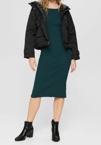 Veste noire matelassée superposée à une robe midi côtelée verte. Des bottines noires complètent le look, mettant l'accent sur le contraste de textures et de couleurs.