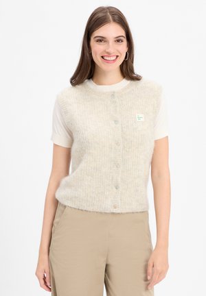 Junge Frau lächelt, trägt eine cremefarbene, knöpfbare, flauschige Weste über einem kurzärmligen weißen Shirt und beigen Hosen vor einem weißen Hintergrund.