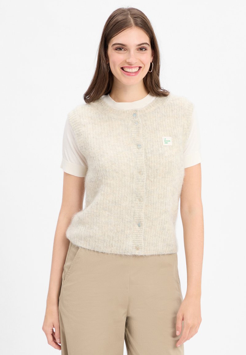 Junge Frau lächelt, trägt eine cremefarbene, knöpfbare, flauschige Weste über einem kurzärmligen weißen Shirt und beigen Hosen vor einem weißen Hintergrund.