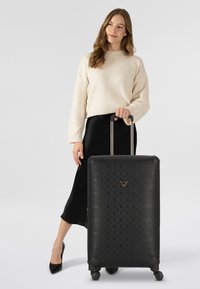 Valigia rigida nera con motivo in rilievo, due manici in metallo e ruote. Accompagnata da una persona con maglione color crema e gonna nera.