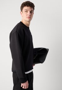 Sudadera negra con puños acanalados y cuello redondo; cuenta con un pequeño logo en la parte delantera. Se sostiene una bolsa de cuero negra y plegada.