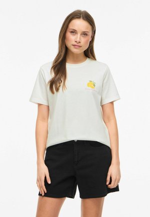 Femme aux longs cheveux bruns portant un t-shirt blanc avec un motif citron jaune et un short noir, debout devant un fond uni.