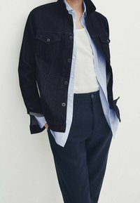 Mörk denimjacka med två bröstfickor, knäppning fram och ståkrage, lager på en ljusblå randig skjorta och mörka byxor.