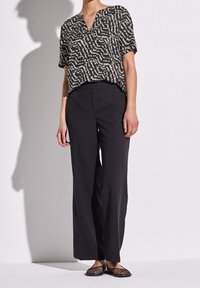 Svarta vida byxor i kombination med en kortärmad topp i v-ringning med ett beige abstrakt mönster, samt platta öppna mesh-skor.