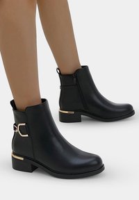 Bottines noires en cuir synthétique lisse, avec un accent doré, une fermeture éclair latérale et un design de talon légèrement empilé.