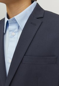 Blazer azul marino con solapa con muesca, tejido texturizado y bolsillo en el pecho, llevado sobre una camisa de cuello azul claro con botones blancos.