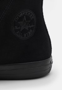 Černá semišová kotníková teniska s texturovaným povrchem, gumovou podrážkou a vyraženým kulatým logem na vnější straně kotníku s nápisem "Converse All Star".