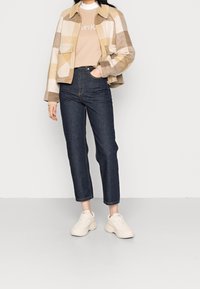 Giacca a quadri beige e marrone con due tasche frontali, abbinata a jeans a vita alta blu scuro e sneakers bianche. Tessuto liscio, forma strutturata.
