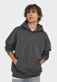 Joven con cabello rizado llevando una sudadera XXL gris oscuro, con las manos en el bolsillo frontal, mirando ligeramente hacia arriba contra un fondo liso.