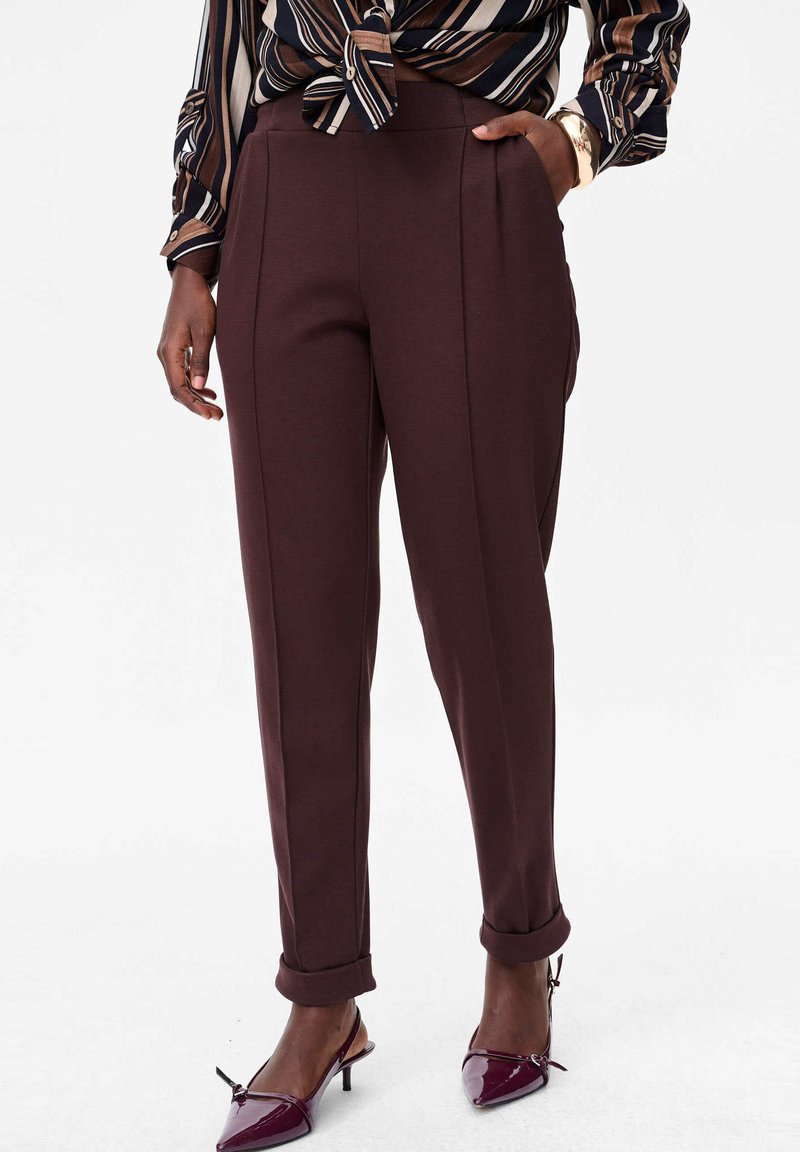 Pantalons marron sur mesure avec une texture lisse, dotés d'une taille haute, de poches latérales et de bas de pantalon retroussés. Associés à des chaussures à talons avec lanières.