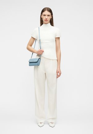 Femme portant un haut blanc à manches courtes en maille côtelée et un pantalon à jambes larges, tenant un petit sac bandoulière bleu sur un fond blanc uni.