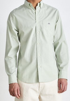 Homme portant une chemise boutonnée à manches longues vert clair avec une poche poitrine et un pantalon de couleur crème, debout devant un fond uni.