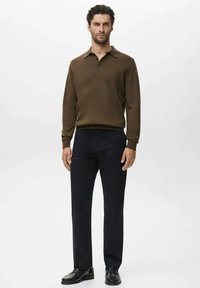 Hombre de pie con suéter de polo de manga larga marrón, pantalones negros y mocasines de cuero negros frente a un fondo claro y liso.