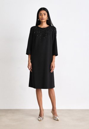 Black knee-length dress with short sleeves, featuring bead detailing on shoulders. Smooth fabric and a loose, straight cut. Worn with silver heels.