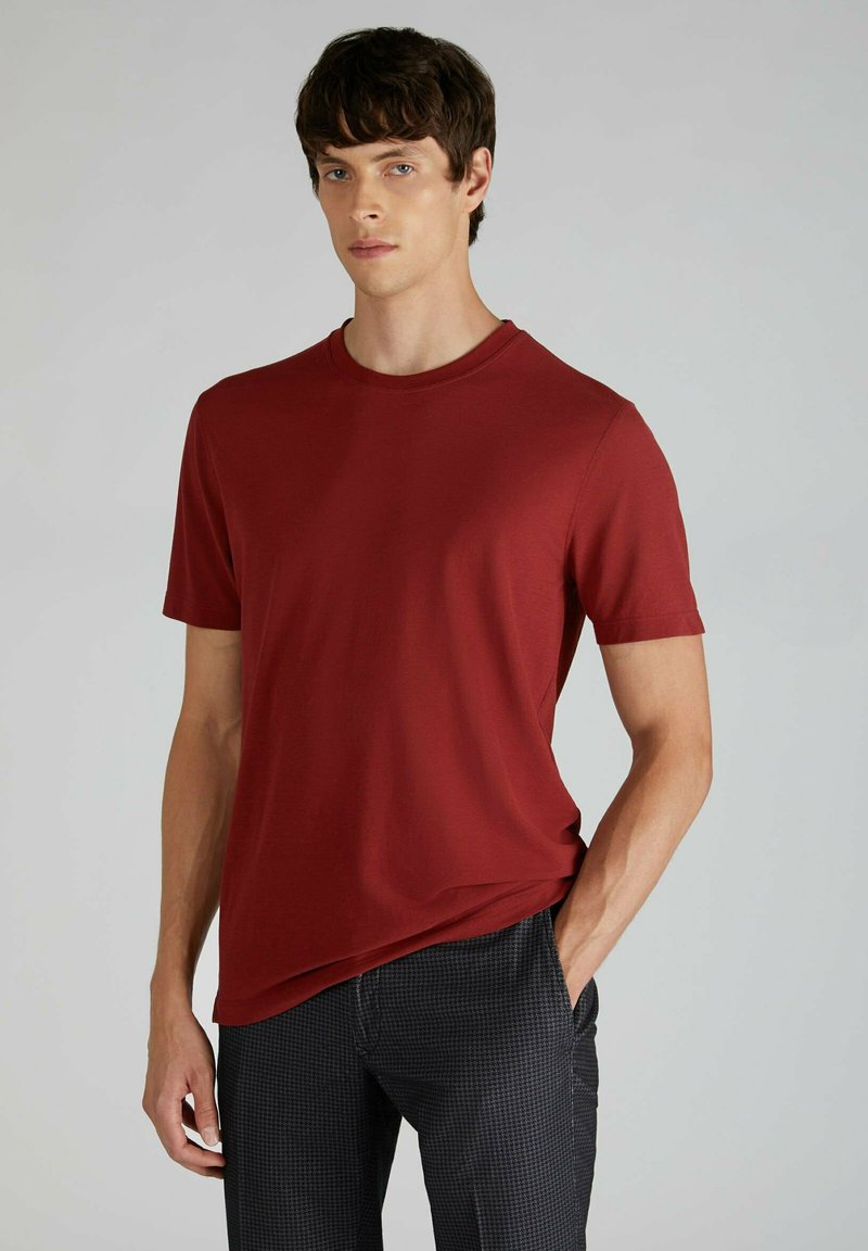 Maglietta rossa a maniche corte con scollatura a girocollo, realizzata in morbido cotone, abbinata a pantaloni neri a fantasia, mani nelle tasche.