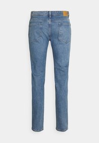 Blå denimjeans med rak skärning, fem fickor och en tanfärgad läderlapp på midjebandet. Klassiska sömnaddetaljer är synliga.