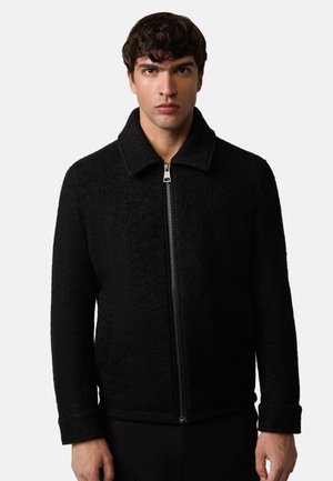 Man wearing a black textured zip-up jacket with a collar, standing against a plain light background, facing forward.