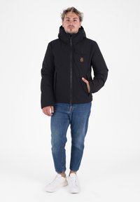 Zwarte gewatteerde jas met een capuchon, vooraan met ritssluiting, zijzakken en een logo-opdruk op de borst, gecombineerd met blauwe spijkerbroek en witte sneakers.