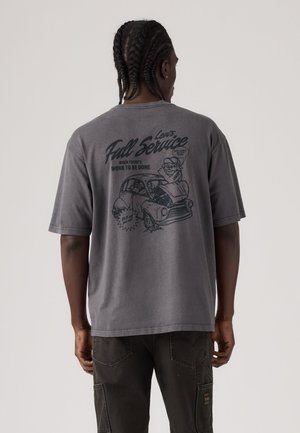 Homme aux cheveux tressés porte un t-shirt gris avec un dessin vintage noir d'un mécanicien réparant une voiture et le texte "Lewis Full Service" au dos.