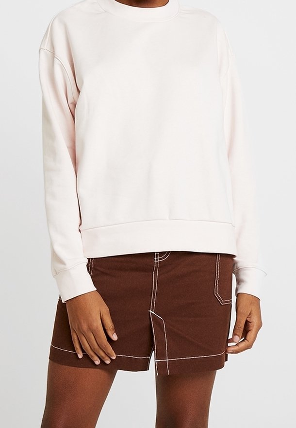 Personne portant un sweat-shirt ample rose pâle et un short marron avec des surpiqûres blanches visibles, debout devant un fond clair et uni.