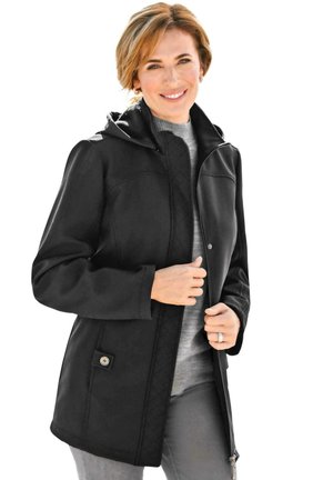 Schwarze wasserdichte Jacke mit Kapuze, ausgestattet mit einer gesteppten Fütterung, Druckknöpfen und seitlichen Taschen. Getragen über einem grauen Pullover und einer Hose.