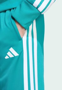 Turkusowe spodnie sportowe z białymi trzema pasami. Posiadają żebrowany pas w talii oraz kieszeń boczną zintegrowaną z designem.