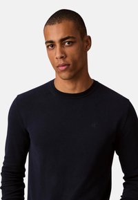 Suéter de cuello redondo azul marino hecho de tela suave con mangas largas y un logo sutil en el pecho. Cuenta con cuello y puños acanalados.