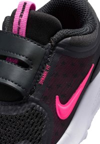 Zapatilla deportiva negra con parte superior de malla, acentos en rosa, etiqueta "DYNAMIC FIT" y una suela blanca contorneada. Presenta una correa en la parte frontal.