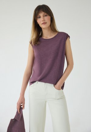 Femme aux cheveux lisses châtain clair portant un haut violet sans manches et un pantalon blanc, tenant une veste violette dans une main.