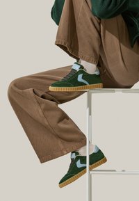 Sneakers in camoscio verde con accenti azzurri chiari, lacci neri e suola in gomma testurizzata. Indossati con pantaloni in velluto a coste marroni e calze bianche.