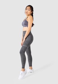 Brassière de sport grise et leggings gris foncé texturés, tous deux ajustés. Chaussures de sport blanches. Modèle debout de profil sur un fond clair.