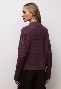 Maglione viola lavorato a maglia con motivo a zigzag testurizzato, caratterizzato da un collo alto e spalle scese, abbinato a pantaloni marrone scuro.