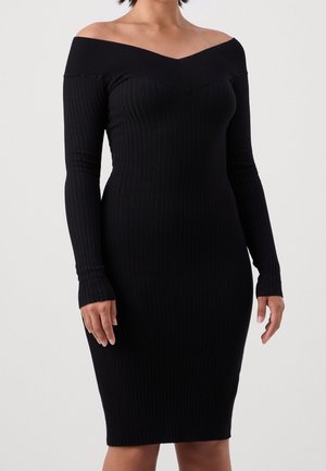 Woman wearing a fitted black ribbed off-shoulder long-sleeve dress, standing against a plain light background.