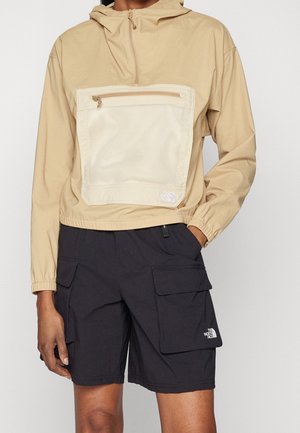 Person trägt eine beige Halb-Reißverschluss-Kapuzenjacke mit einer großen vorderen Netz-Tasche und schwarze Cargo-Shorts mit Klappentaschen.