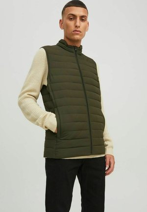 Vesta verde cu umplutură, cu guler înalt și închidere cu fermoar, asociată cu un top crem texturat cu mâneci lungi. Îmbrăcămintea include pantaloni negri.