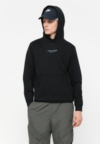 Sort hættetrøje med en frontlomme, prydet med blå tekst "WEEKEND OFFENDER ADMIT NOTHING." Sammen med en mørk kasket, skaber det et monokromt look.