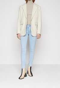 Blazer beige sobre un jersey de cuello alto beige, combinado con jeans ajustados azul claro y botines bicolor con acentos en negro y beige.