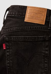 Poche arrière d’un jean en denim noir avec patch en cuir de la marque Levi Strauss et étiquette en tissu rouge sur la couture latérale.