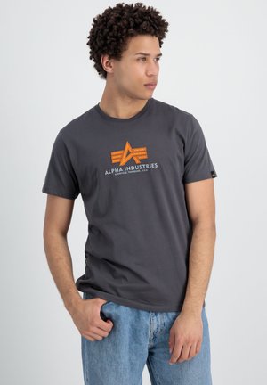 Camiseta de algodón gris con un logo gráfico en naranja y blanco que dice "ALPHA INDUSTRIES" en el frontal. Cuello redondo y mangas cortas, corte relajado.