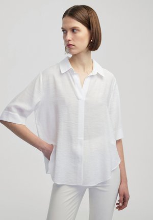 Witte, kortarmige blouse met een knoopsluiting en V-hals. Licht, gestructureerde stof met een comfortabele pasvorm. Heeft een subtiele borstzak.