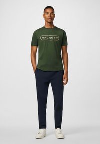 Camiseta de algodón verde con el logo "HACKETT" en beige, combinada con pantalones de chándal azul marino y zapatillas blancas; ajuste casual, mangas cortas, diseño sencillo.