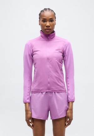 Žena oblečená v priliehavom levanduľovom zipovacom tričku a zodpovedajúcich krátkych nohaviciach, stojaca pred jednoduchým bielym pozadím.