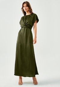 Maksi obleka v temno zeleni satenski tkanini s zasukanim dizajnom v pasu, rokavi na kapico in gladko teksturo. Obuta v sandale bež barve s trakovi.