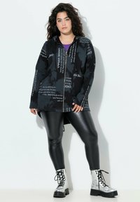 Sudadera negra con cremallera y patrones de texto, combinada con leggings negros brillantes y botas de combate plateadas con cordones. El diseño presenta un corte holgado y bolsillos.