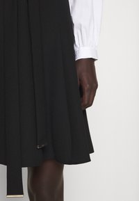 Zwarte geplooide rok met een gladde textuur, voorzien van een zijstrikceintuur met een metalen accent. Gestyled met een witte blouse met lange mouwen.